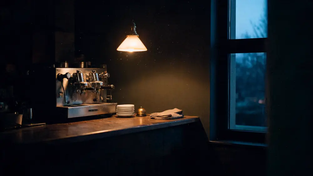 The counter of a small cafe after closing: an espresso machine wiped down, a stack of saucers and a brass bell under one pendant lamp.