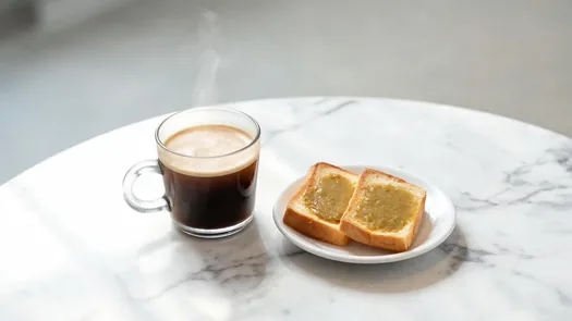 A glass of kopi and a plate of kaya toast on a marble table.